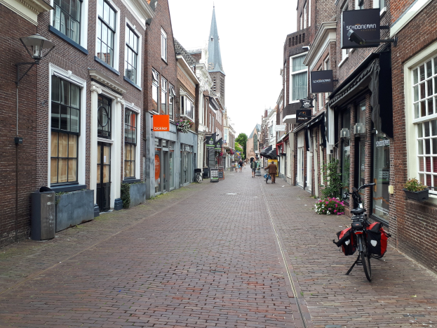 Ein Fahrrad, das auf einer städtischen Straße in Delft, Niederlande, geparkt ist und von Gebäuden mit Fenstern, Blumentöpfen und einem Müllcontainer flankiert wird.