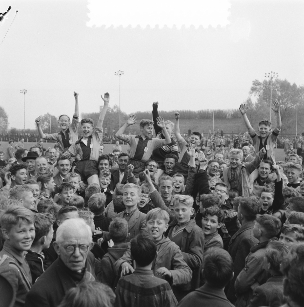 Schwarzes und weißes Foto einer Menge vor einem Stadion, einige halten Hände in die Luft, mit Laternenmasten, Bäumen und einem klaren Himmel im Hintergrund.