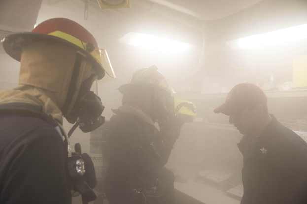 Feuerwehrleute in einem raucherfüllten Raum während einer Übung, einer hält eine Kamera, trägt Helme und Schutzausrüstung.