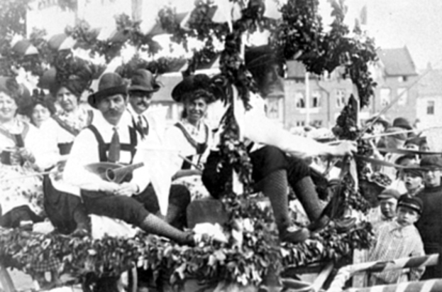 Eine Gruppe von Menschen in traditioneller bayrischer Tracht, einige mit Musikinstrumenten, fährt auf einemlaubgeschmückten Festwagen bei einem Umzug mit, im Hintergrund sind Gebäude und ein klarer Himmel zu sehen, dargestellt in Schwarz-Weiß.