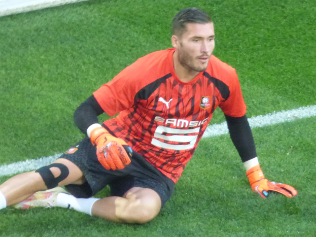Ein Fussballspieler sitzt auf dem Boden mit Händen auf Knien, trägt ein rotes T-Shirt, schwarze Shorts, weisse Socken und orange Baseball-Handschuhe.