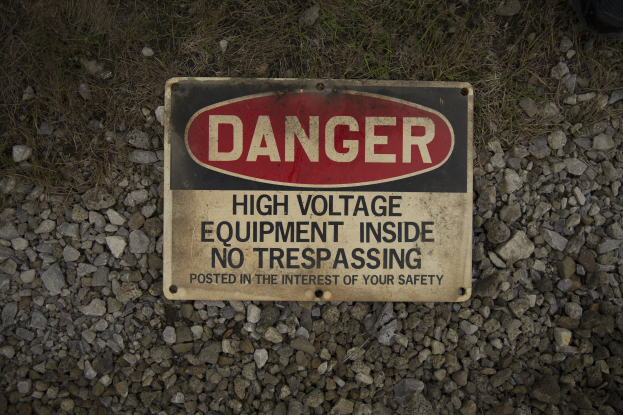 Eine Gefahrenzeichen auf dem Boden umgeben von Steinen und Gras, auf dem steht "Hochspannungsgerät Inside No Trespassing".