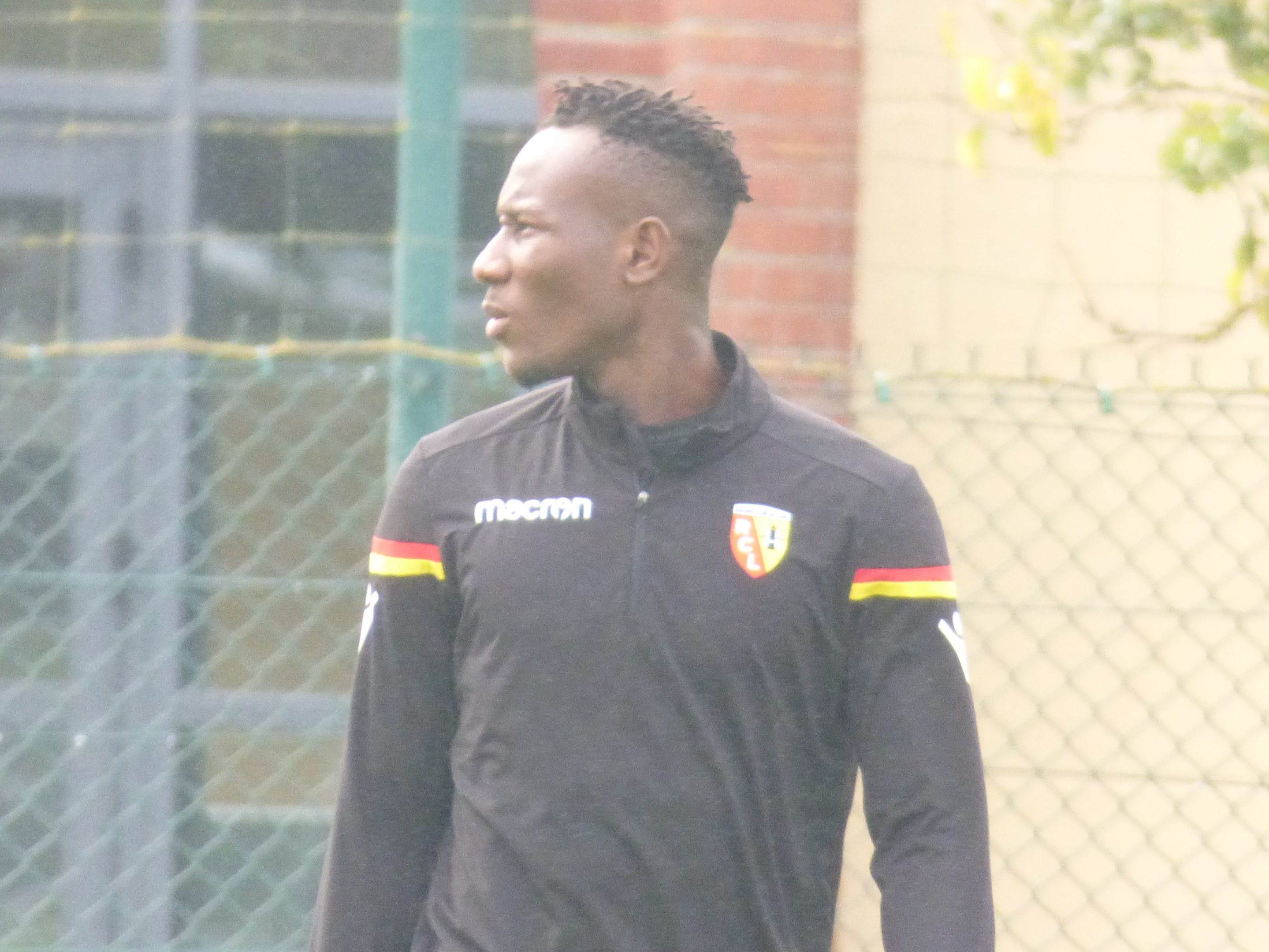 Ein Mann in einer schwarzen Jacke steht auf einem Fußballfeld, umgeben von einem Zaun und einem Baum, und sieht konzentriert während eines Trainingssitzung aus.