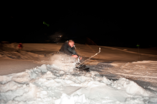 Eine Person beim Skifahren auf Schnee mit einer weiteren Person im dunklen Hintergrund.