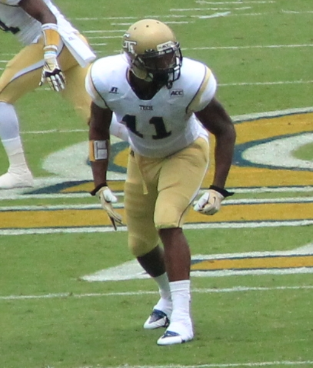 A football player in a sports uniform, helmet, and cleats runs with the ball on a field.