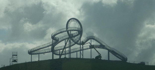 Eine Achterbahn auf dem Gipfel eines Hügels unter einem bewölkten Himmel, umgeben von Stützpfählen und -strukturen.