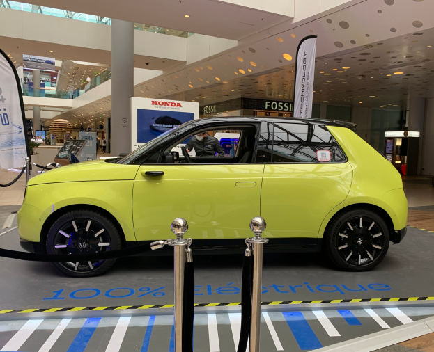 Ein gelbes Wasserstoff-elektrisches Auto vor einem Gebäude mit Absperrpfosten, Bannern, Menschen, Schildern, einer Topfpflanze, Deckenleuchten und Lüftern.