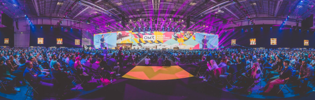 Eine große Menschenmenge sitzt vor einer hell erleuchteten Bühne mit mehreren stehenden Personen, wahrscheinlich auf einer Online-Marketing-Rockstars-Veranstaltung.