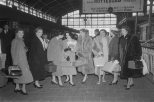 Schwarz-weißes Bild einer Gruppe von Frauen, die zusammen an einem Bahnhof stehen, jede hält eine Tasche in der Hand, mit einem Zug auf der linken Seite und Geländern und einer Tafel auf der rechten Seite.