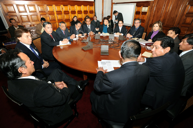 Eine Runde Tischbesprechung mit Delegierten, Mikrofonen, Papieren, Kabeln und Dateien auf dem Tisch, mit einer hölzernen Wand im Hintergrund.