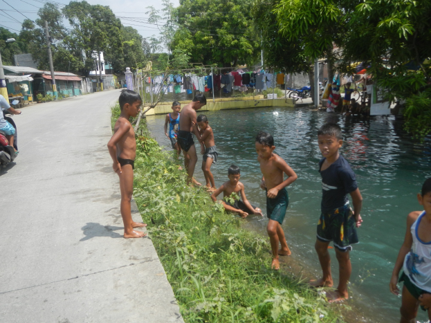 Gruppe von Kindern, die in flachem Straßenwasser spielen, einige stehen, während andere auf dem Gras sitzen, mit einer Person auf einem Motorrad links und Häusern, Bäumen und Strommasten im Hintergrund unter einem klaren blauen Himmel.