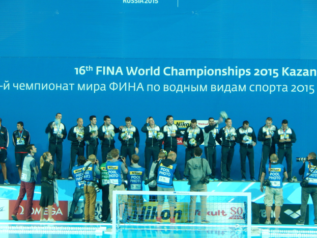 Gruppe von Menschen auf einem Schwimmbeckenrand mit Kameras, einem Netz im Vordergrund und einem Banner mit der Aufschrift "FINA World Championships 2015 Kazan" im Hintergrund.