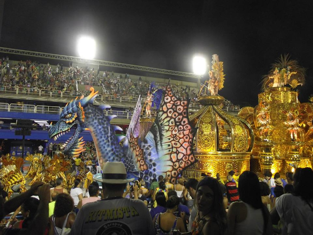 Eine große Menschenmenge um einen hell erleuchteten, bunten Festwagen herumstehend, mit einem Geländer um den Festwagen und einem Gebäude im Hintergrund unter einem dunklen Himmel.