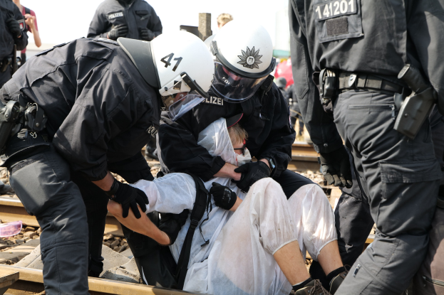 Eine Gruppe von Polizisten in Einsatzausrüstung nimmt einen Mann in einem weißem Kleid auf einem Bahngleis fest, mit anderen im Hintergrund sichtbar.