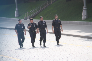 Drei Polizisten in Uniform und Mützen gehen eine Straße entlang mit Pfählen, Geländern, Treppen und Gras im Hintergrund.