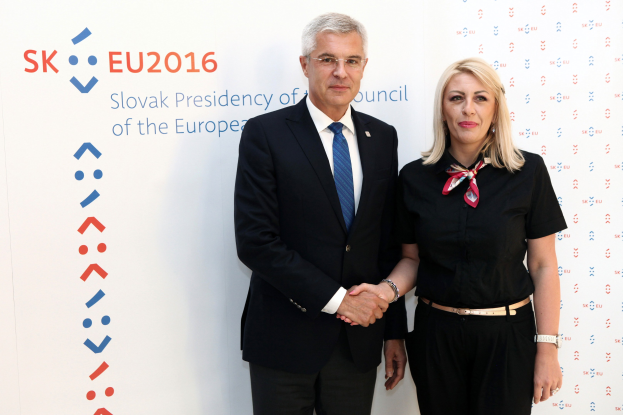 Zwei Personen, ein Mann in einem schwarzen Blazer und weißem Hemd mit einer blauen Krawatte und eine Frau mit Brille, stehen lachend vor einer weißen Tafel mit der Aufschrift "sk eu 2016", identifiziert als der Präsident des Europäischen Rates.