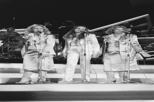 Drei Frauen singen in Mikrofone auf der Bühne mit Musikern hinter ihnen, schwarz-weiß.