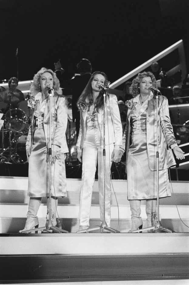 Drei Frauen singen in Mikrofone auf der Bühne mit Musikern hinter ihnen, schwarz-weiß.