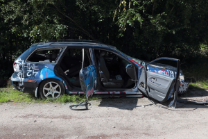 Ein geschädigtes Auto am Straßenrand mit Gras und Bäumen im Hintergrund.