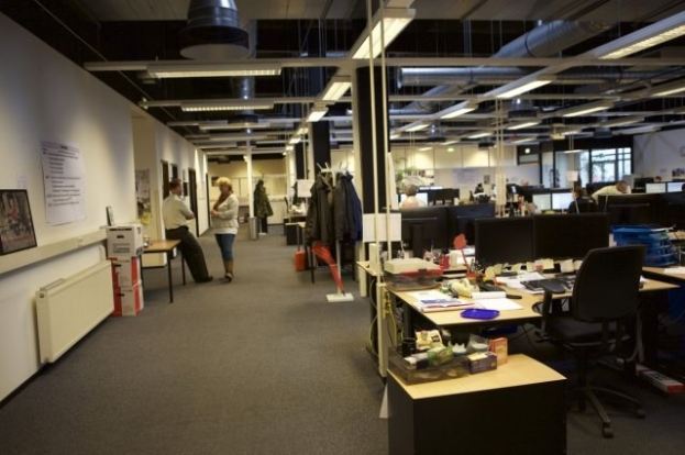 Großes Großraumbüro mit Menschen an Schreibtischen, Computern und Monitoren, umgeben von Papieren, Büchern, Kartons und Wandtafeln mit Text und Bildern, unter Deckenleuchten mit Glasfenstern im Hintergrund.