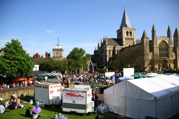 Ein historisches Gebäude mit Bäumen davor, zusätzliche Gebäude und ein Turm im Hintergrund, Menschen auf einem Weg mit Geschäften und andere auf der nahen Wiese sitzend.