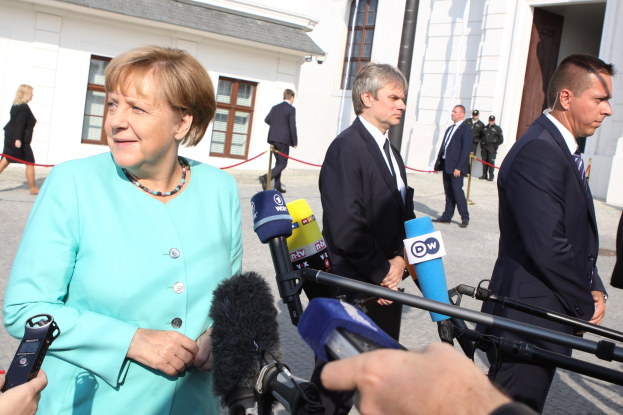 Bundeskanzlerin Angela Merkel spricht vor der Reichskanzlei in Berlin mit Reportern, umgeben von einer Gruppe von Menschen, einige halten Mikrofone, mit Gebäuden im Hintergrund.