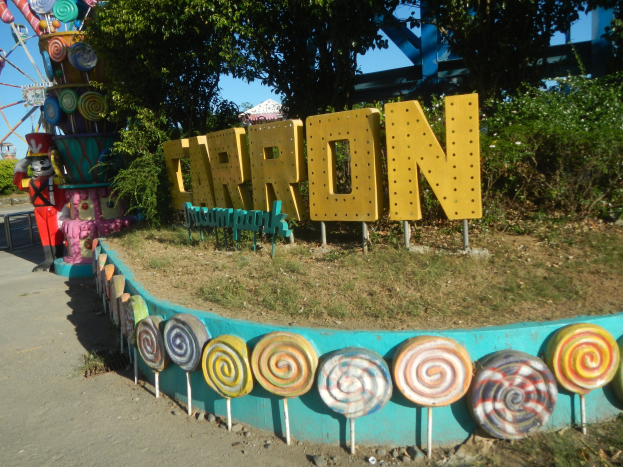 Eine Karussellfahrt mit einem riesigen Riesenrad im Hintergrund, umgeben von Bäumen und Gras, mit einer Person auf der linken Seite, einer Straße unten und Lollis und einem Namensschild im Vordergrund.