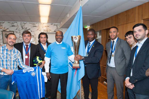 Eine Gruppe von Menschen, die zusammenstehen, wobei zwei in der Mitte einen Pokal halten, einer links ein T-Shirt und ein anderer links einen Ball, sowie eine Flagge im Hintergrund.