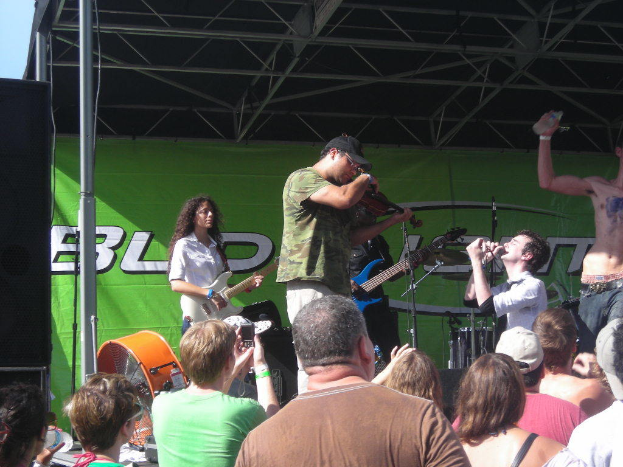 Eine Gruppe von Musikern, die auf der Buhne verschiedene Instrumente spielen, waehrend das Publikum vor ihnen steht und die Musik geniesst, mit einem grossen schwarzen Lautsprecher links und einem gruenen Banner im Hintergrund.