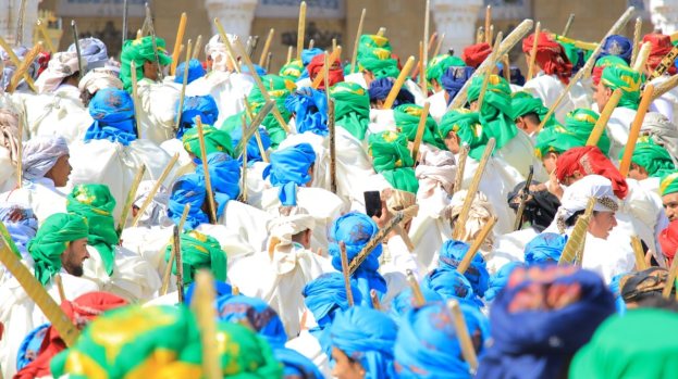 Eine große Gruppe von Menschen in bunten Kostümen marschiert die Straße entlang, jeder hält einen Stock in der Hand, während ein Festival mit einem Gebäude im Hintergrund stattfindet.