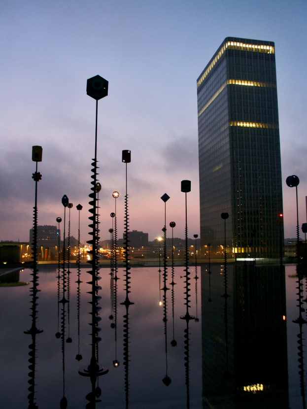Mehrere Pole und Gebäude mit Lichtern unter einem bewölkten Himmel, mit Spiegelungen der Pole