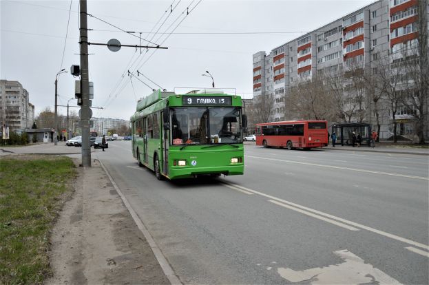 Grüner Bus fährt auf einer Straße mit hohen Gebäuden, Gras auf der linken Seite, Strommasten mit Drähten, Bäumen im Hintergrund und einem klaren blauen Himmel.