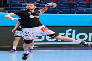 Ein Mann in schwarzem Hemd und weißen Shorts hält einen Handball und bereitet sich auf das Spiel vor, mit Zuschauern im Hintergrund eines Stadions.