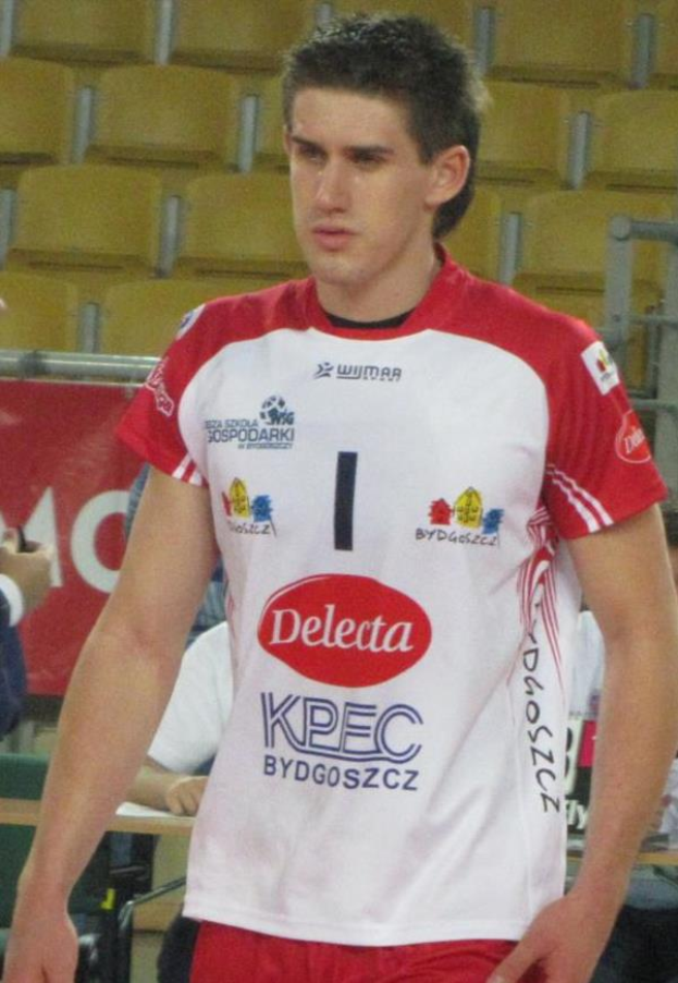 Ein Mann in roter und weißer Handballdress steht auf einem Platz, entschlossen schauend, mit Zuschauern und einer Flagge mit der Aufschrift "Krasnoyarsk vs Bydgoszcz" im Hintergrund.