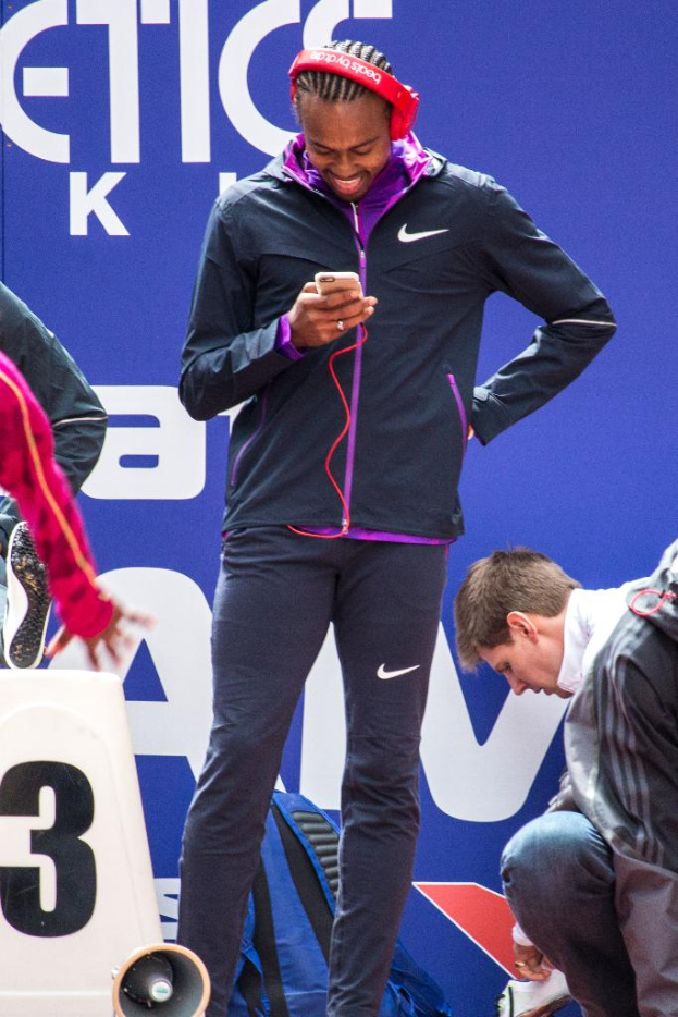 Eine Person, die ein Mobiltelefon hält und strahlt, mit einer weiteren Person rechts im Schneidersitz, einem Rucksack, einem Megafon unten, einer Wand im Hintergrund und einer weiteren Person links stehend.