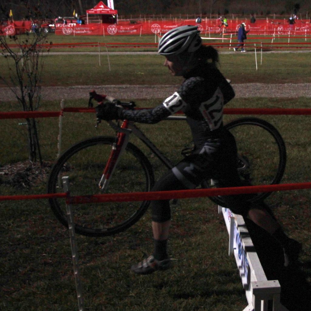Eine Frau in einem schwarzen Kleid und Helm hält ein Fahrrad, geht auf Gras neben einem Zaun mit einem roten Tuch, mit Menschen und Zelten im Hintergrund.