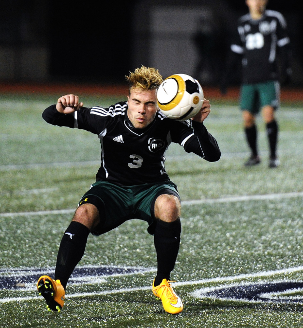 Ein Mann in schwarzem T-Shirt, grünen Shorts, gelben Schuhen und schwarzen Socken spielt Basketball auf einem Court und hält einen weißen Ball, während ein anderer Mann im Hintergrund zusieht.