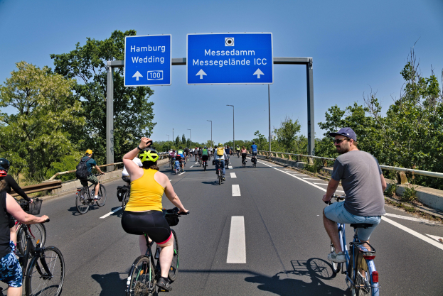 Gruppe von Radfahrern in Helmen auf einer Straße mit Bäumen auf einer Seite und Laternen im Hintergrund unter einem klaren blauen Himmel, mit einem Hamburger Radtour-Schild oben.