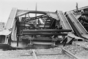 Ein Schwarz-Weiß-Foto einer Bahnschiene, die zu einer baufälligen Brücke mit zerbrochenen Geländern und Schutt führt, umgeben von Gebäuden unter einem klaren Himmel.