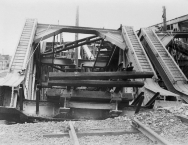 Ein Schwarz-Weiß-Foto einer Bahnschiene, die zu einer baufälligen Brücke mit zerbrochenen Geländern und Schutt führt, umgeben von Gebäuden unter einem klaren Himmel.