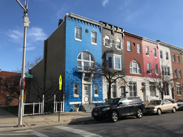 Eine Reihe bunter Häuser an einer Straßenkreuzung in Baltimore, Maryland, mit parkenden Autos, Bäumen, Strommasten, einem Zaun und einem bewölkten Himmel.