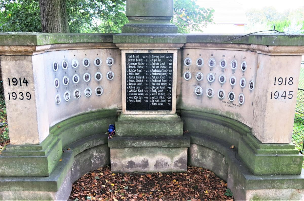 Ein Denkmal in einem von Bäumen und einem Zaun umgebenen Park mit einer Tafel an der Wand und trockenen Blättern auf dem Boden, das sich auf dem jüdischen Friedhof in Berlin befindet und als Gedenkstätte für Holocaust-Opfer dient.