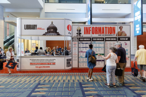 Eine Gruppe von Menschen auf dem Nationalen Buchfest, einige sitzen und einige stehen, vor einem Stand mit Bannern, einem Anzeigebildschirm, einer Treppe, einem Geländer und einer Decke mit Deckenleuchten.