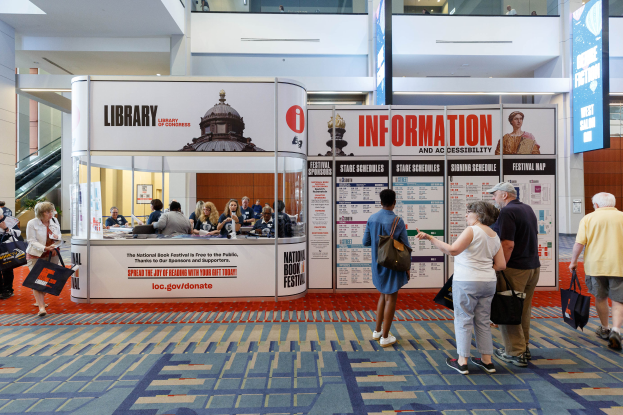 Eine Gruppe von Menschen auf dem Nationalen Buchfest, einige sitzen und einige stehen, vor einem Stand mit Bannern, einem Anzeigebildschirm, einer Treppe, einem Geländer und einer Decke mit Deckenleuchten.