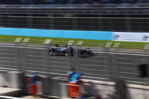 Ein Formel-1-Auto fährt auf einer Rennstrecke vorbei an einer Menge Zuschauern am Straßenrand, Gras auf dem Boden und Schildern im Hintergrund.