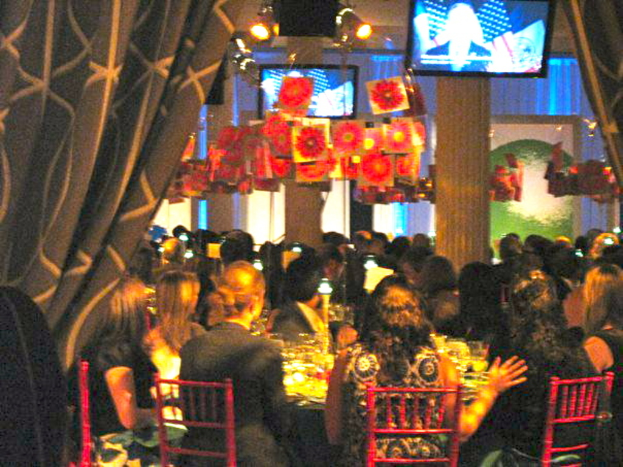 Eine Gruppe von Menschen sitzt an Tischen mit Essen, Fernsehgeräten, Lichtern, hängenden Papieren und Vorhängen in einem Raum.