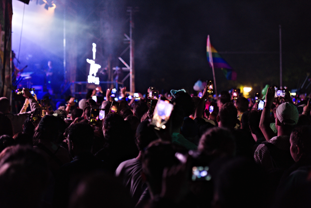 Menschenmenge bei einem Konzert mit hochgehaltenen Handys, beleuchtet von Scheinwerfern, umgeben von Masten und einer Fahne, mit Musikern auf der Bühne.