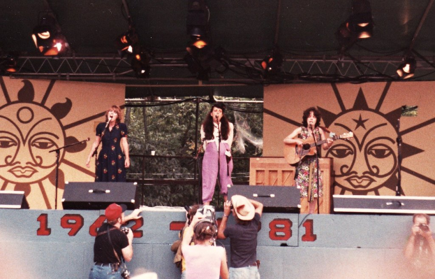 Drei Mädchen auf der Bühne, zwei singen in Mikrofone und eines spielt Gitarre, mit Publikum, das im Vordergrund Fotos macht und einem braunen Bühnenrahmen mit Scheinwerfern im Hintergrund.