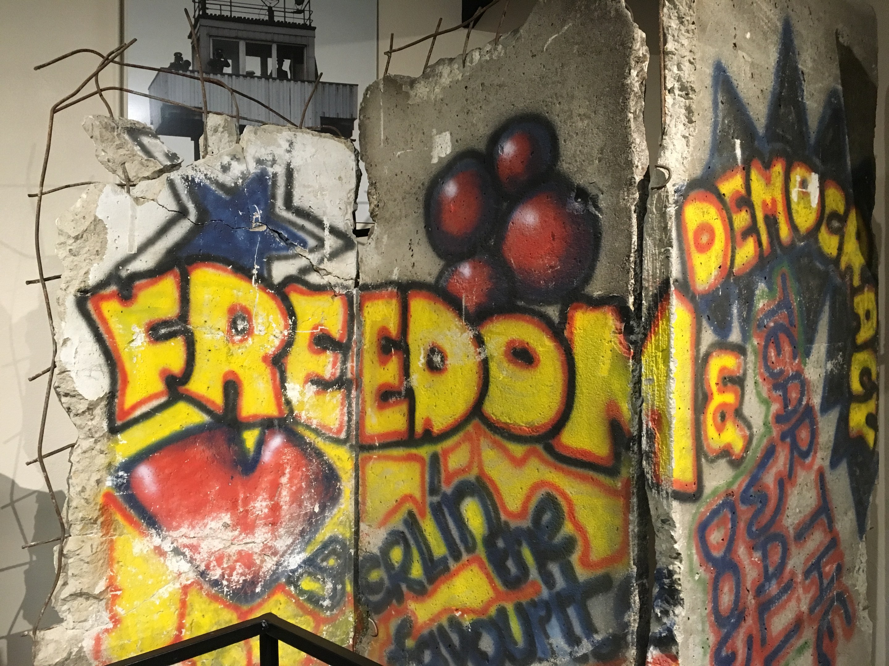 Graffiti an einer Wand mit der Aufschrift 'Freiheit und Demokratie' in fetten schwarzen Buchstaben, mit einem Geländer darunter und einem Gebäude mit Fenstern im Hintergrund.