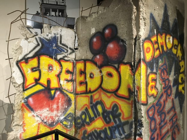 Graffiti an einer Wand mit der Aufschrift 'Freiheit und Demokratie' in fetten schwarzen Buchstaben, mit einem Geländer darunter und einem Gebäude mit Fenstern im Hintergrund.